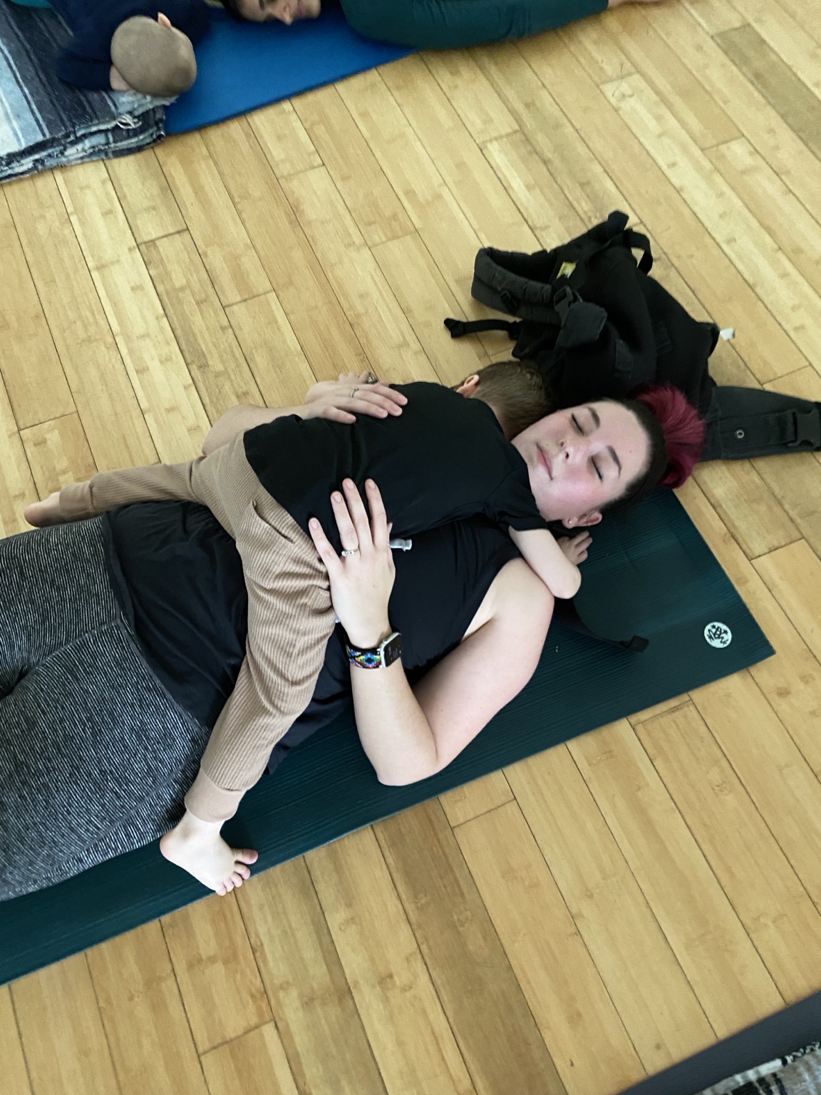 A parent resting on a yoga mat while holding a young child